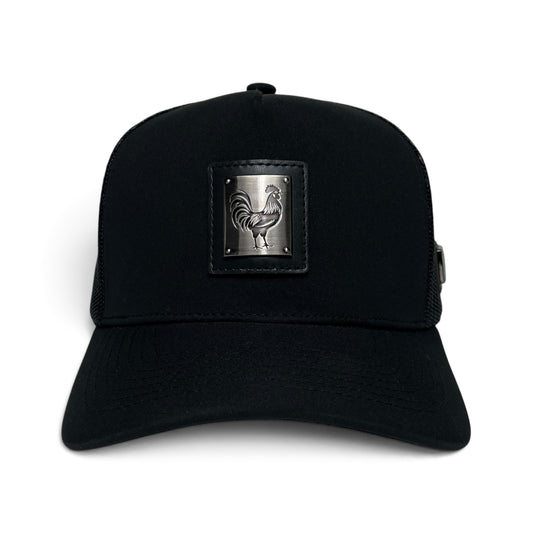 A black trucker hat featuring a rectangular black leather patch on the front with an embossed silver metal rooster design.